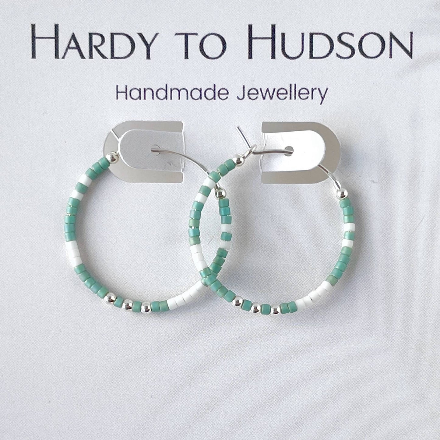 Handmade sterling silver hoop earrings with green and white Miyuki beads and silver accent beads on a beige background, branded as Hardy to Hudson.