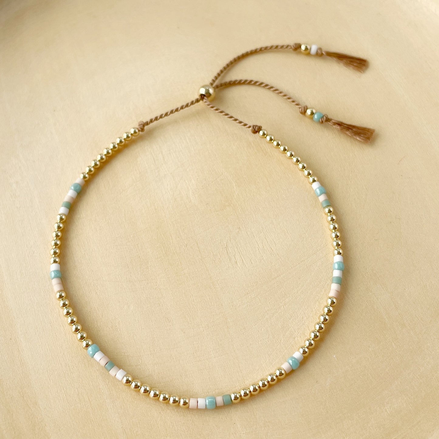 Beige silk bracelet with aqua, blush and white glass beads and 14k gold fill accents on a wood background