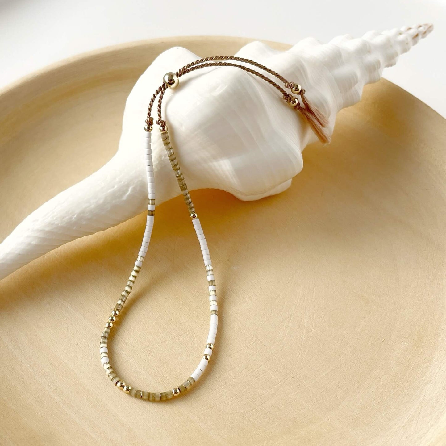 Delicate beaded bracelet with tiny beige and white glass beads and gold fill accents. Displayed against a white shell on a wooden plate