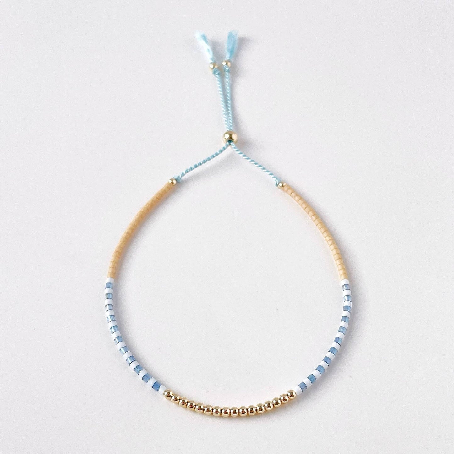 dainty seed bead bracelet with beige, blue and white beads and 14k gold filled accents on pale blue silk cord with a slider bead