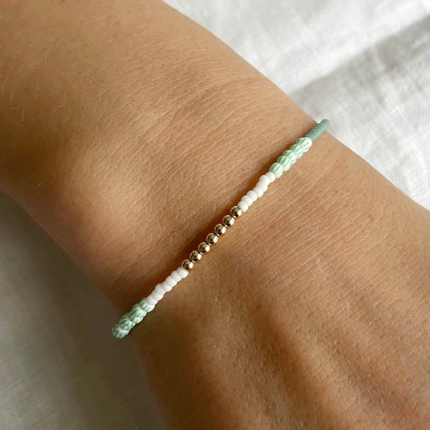 Close-up of a wrist wearing a bracelet with green, white, and gold beads on a light background.