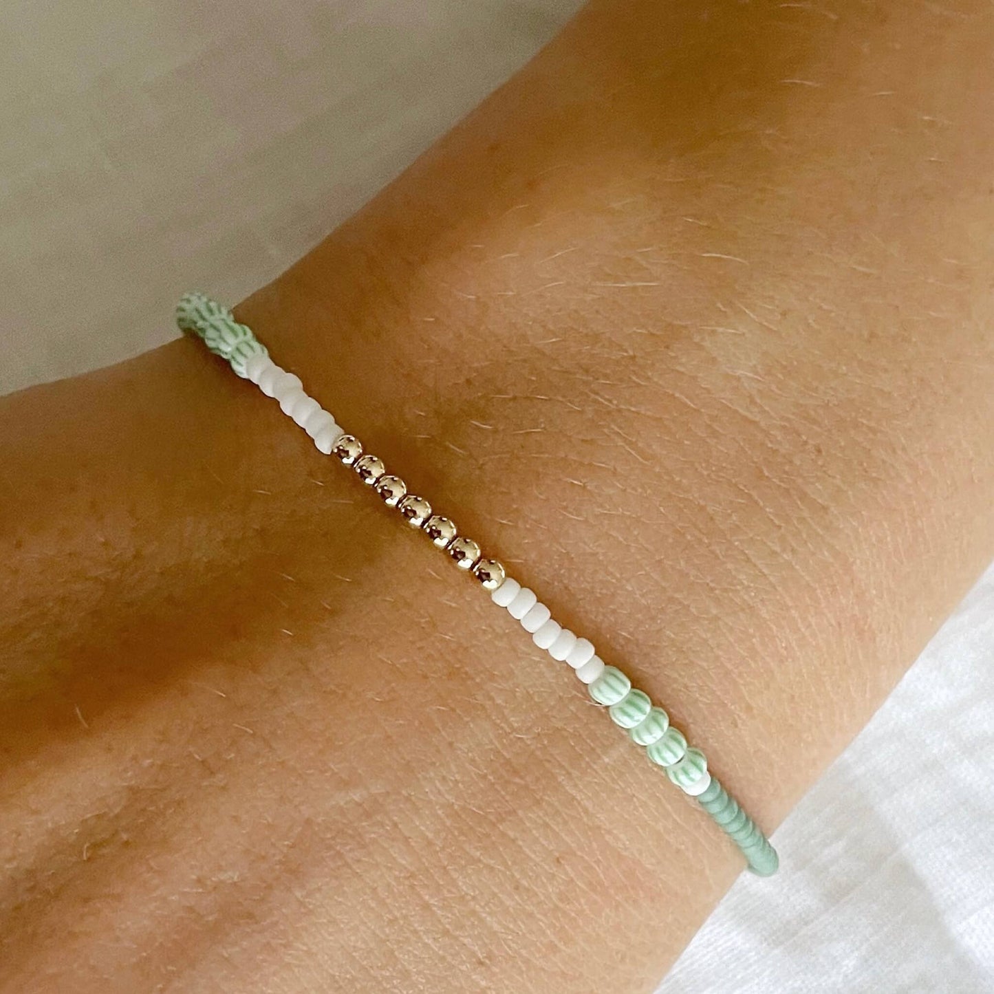 Close-up of a wrist wearing a bracelet with green, white and gold beads on a neutral background