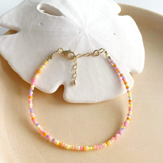 Beaded bracelet with pink, yellow, orange and lilac glass beads and gold fill accents displayed draped over a sand dollar