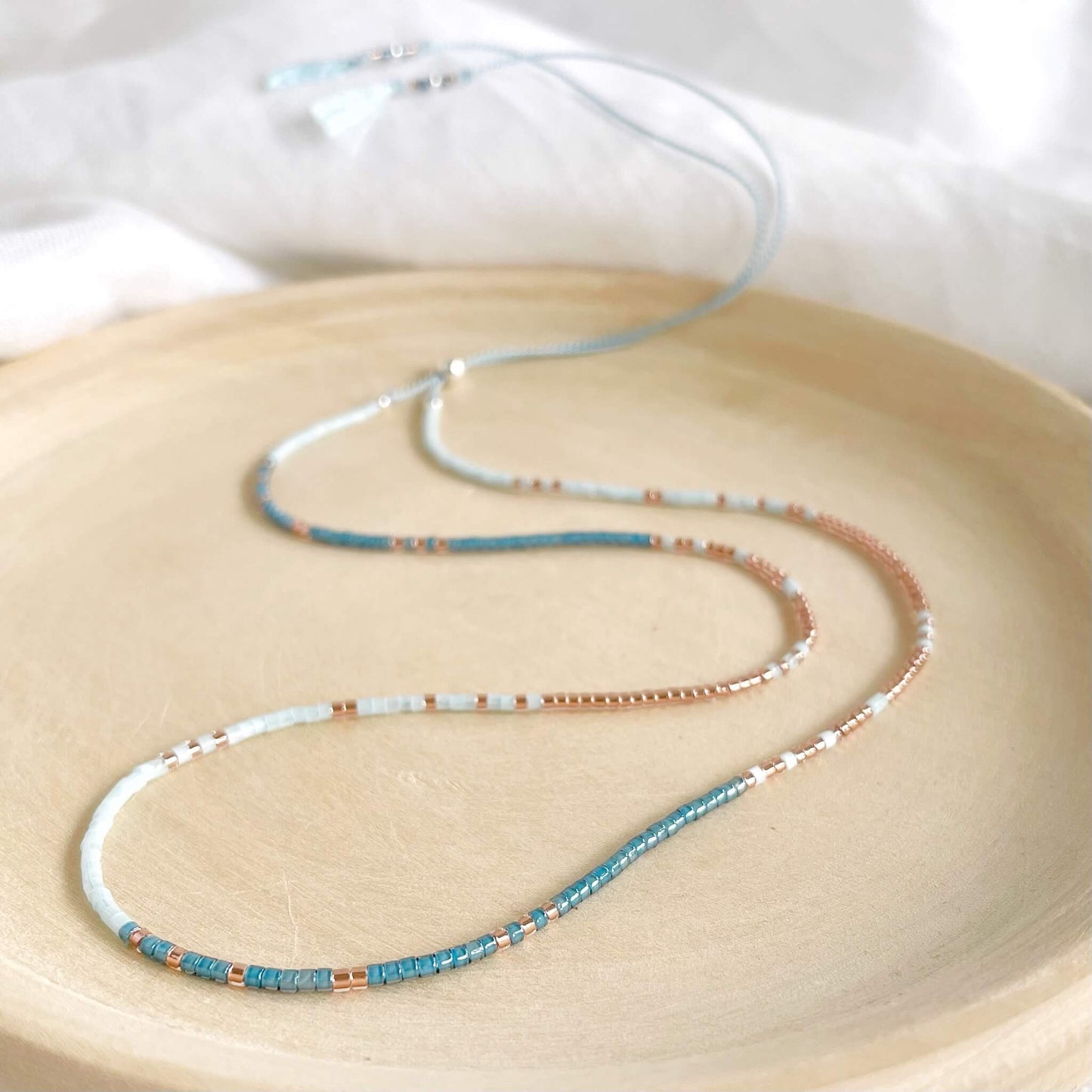 dainty Blue and copper glass beaded necklace on pale blue silk cord with silver clasp displayed on a wooden dish