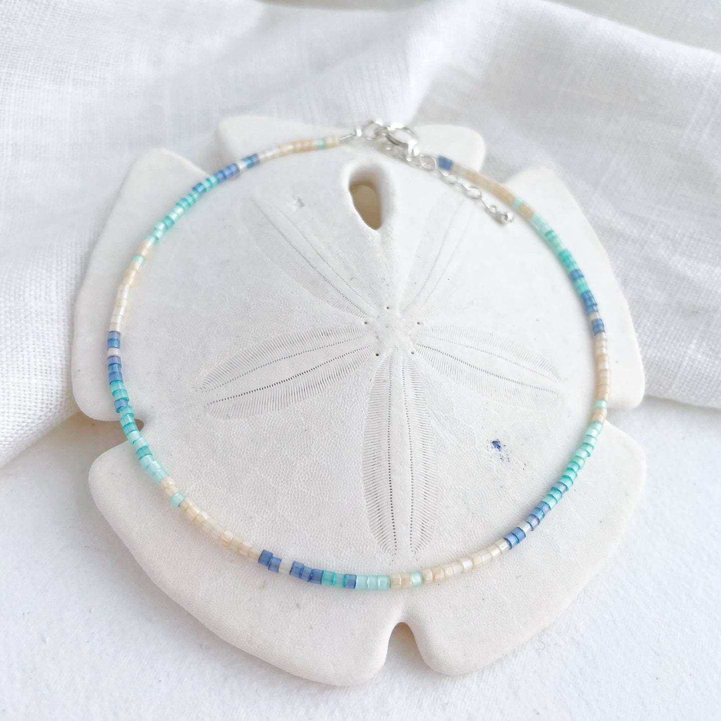Delicate anklet with beige, aqua and blue Japanese glass beads and sterling silver clasp displayed draped on a white sand dollar