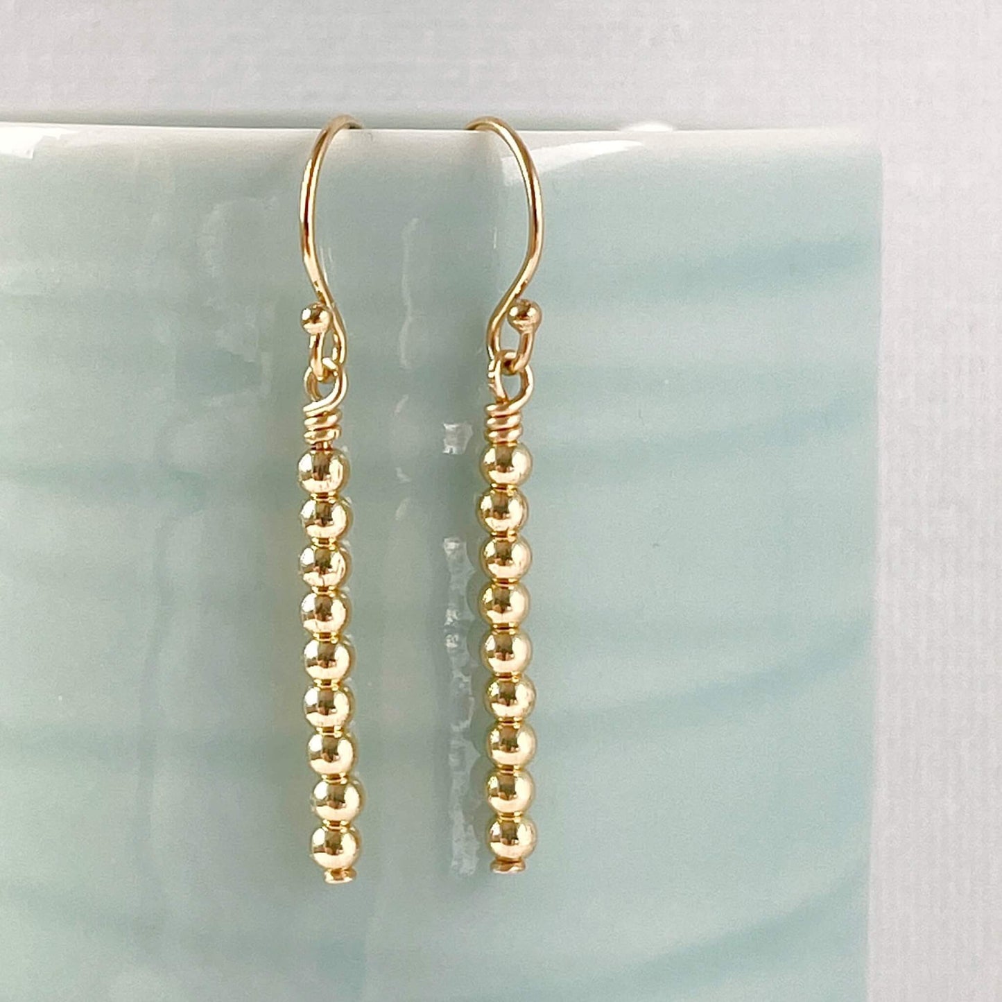 Minimalist Gold beaded dangle earrings on a light aqua surface
