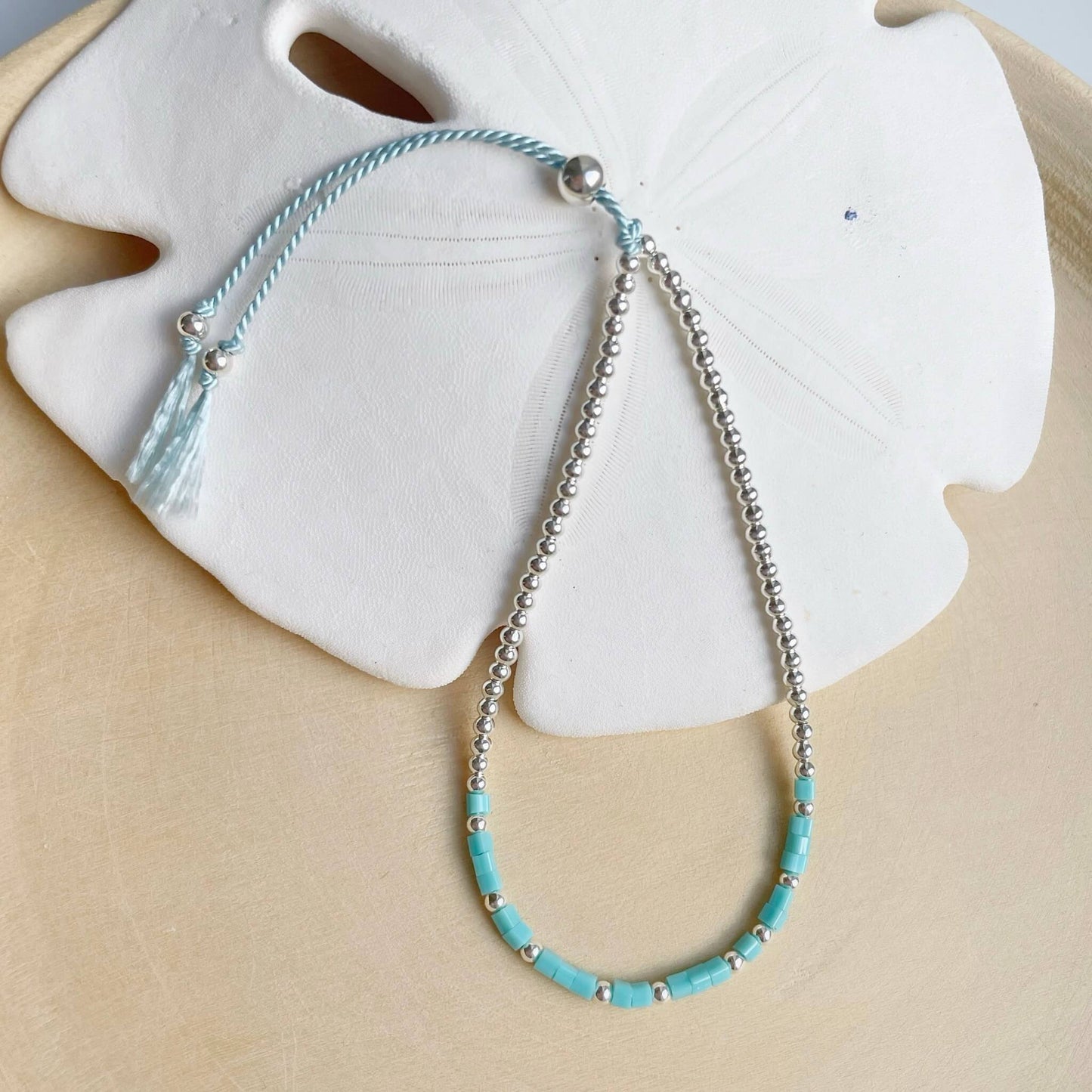 dainty bracelet with turquoise square glass beads and sterling silver 2mm beads on pale blue silk with a sliding clasp displayed draped over a sand dollar