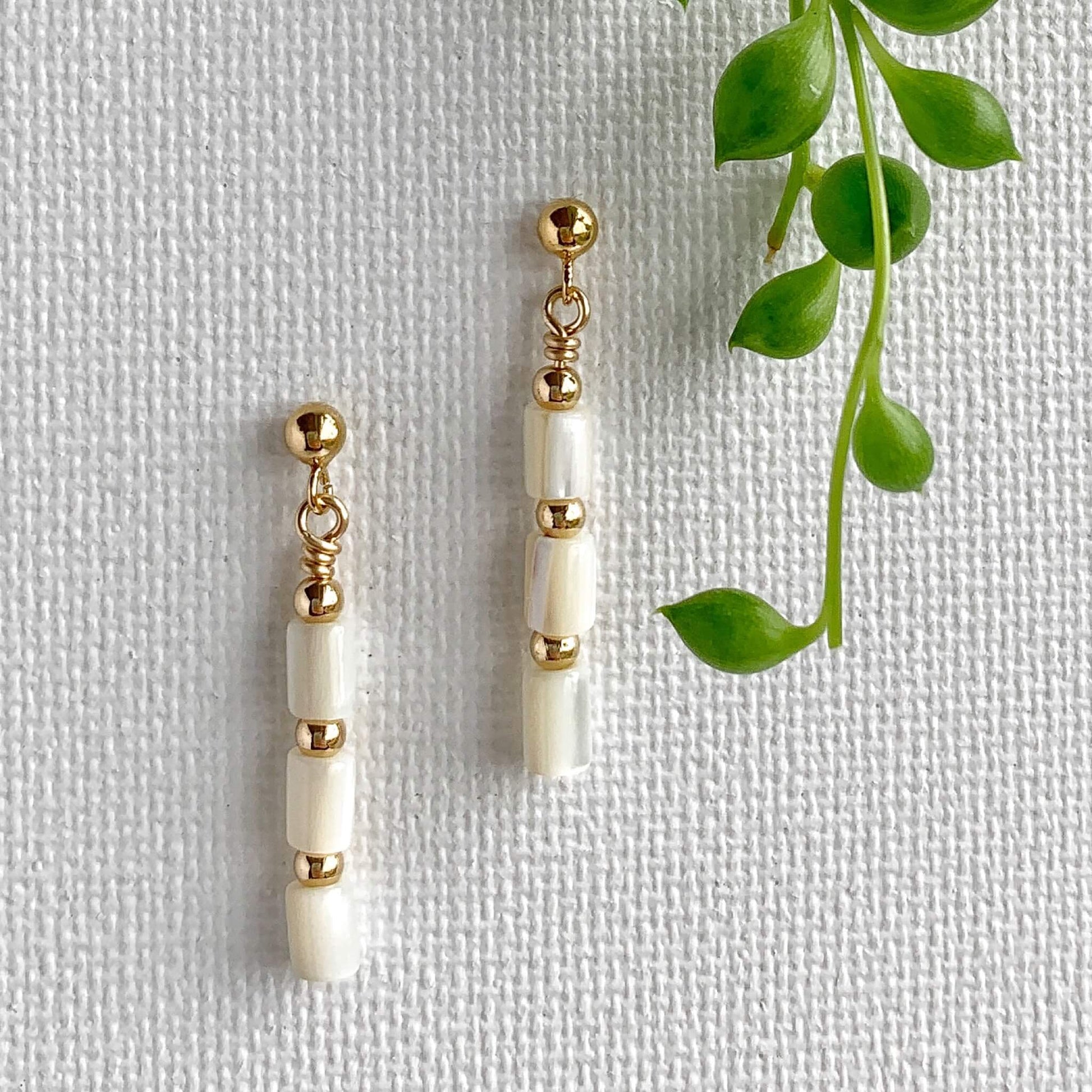 dainty gold stud dangle earrings with mother of pearl cylinder beads against a white background with a succulent plant