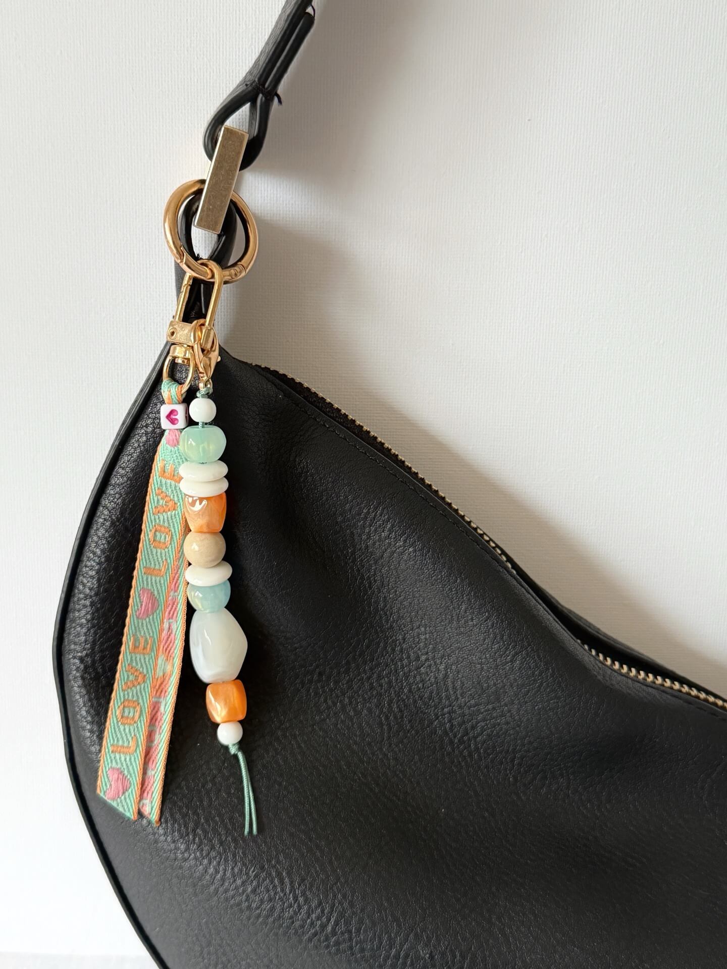 beaded bag charm with a love embroidered ribbon and aqua, white and orange chunky resin beaded on a gold clasp shown attached to a black bag
