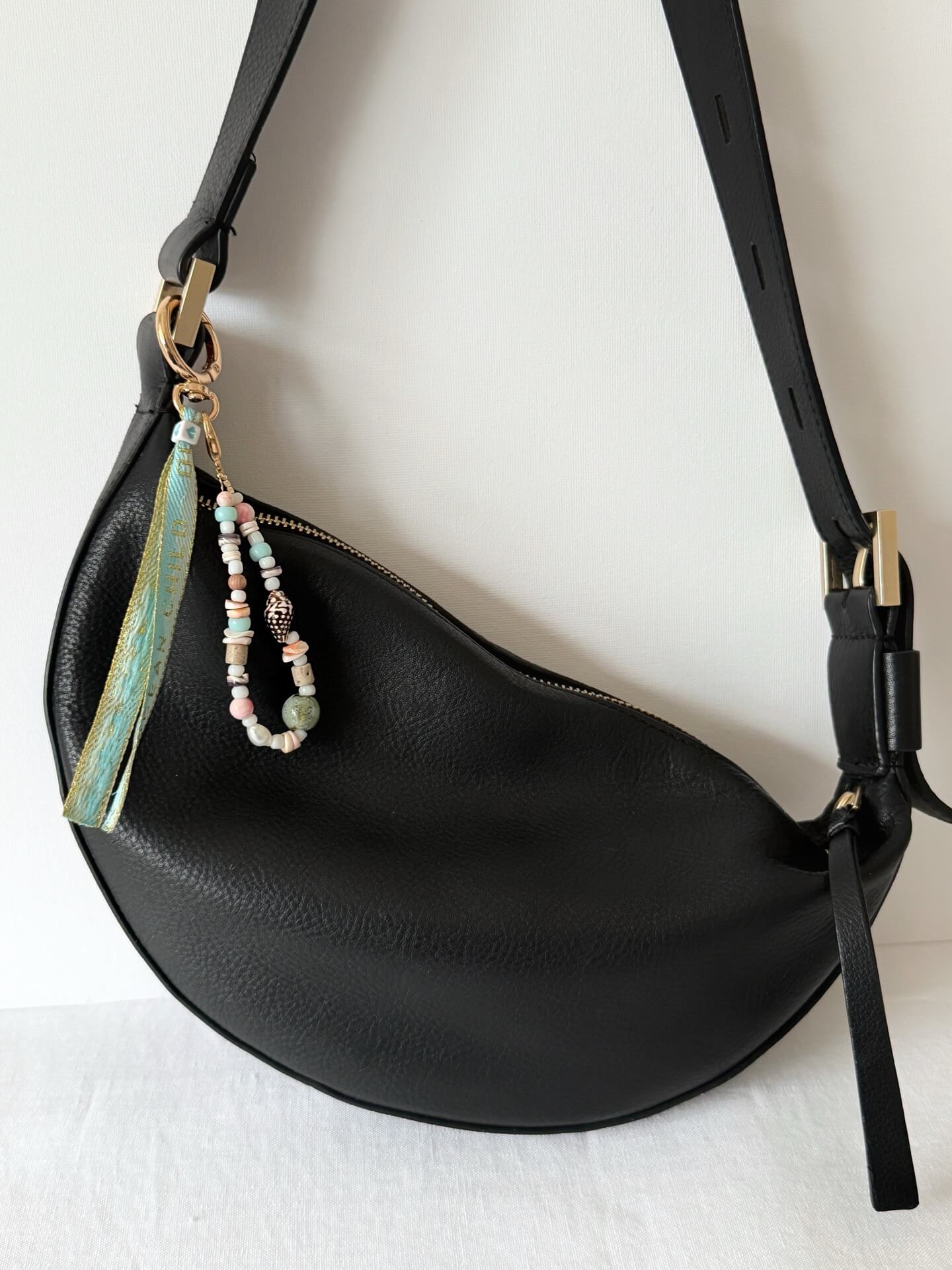 golden carabiner clasp bag charm with shell, glass, and wood beads with a blue and gold embroidered ocean child ribbon tassel attached to a black bag