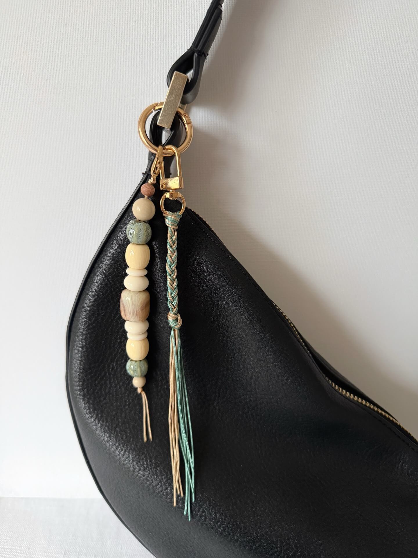 golden carabiner bag charm with blue and beige chunky beads with a braided nylon cord tassel in aqua and beige displayed on a black bag
