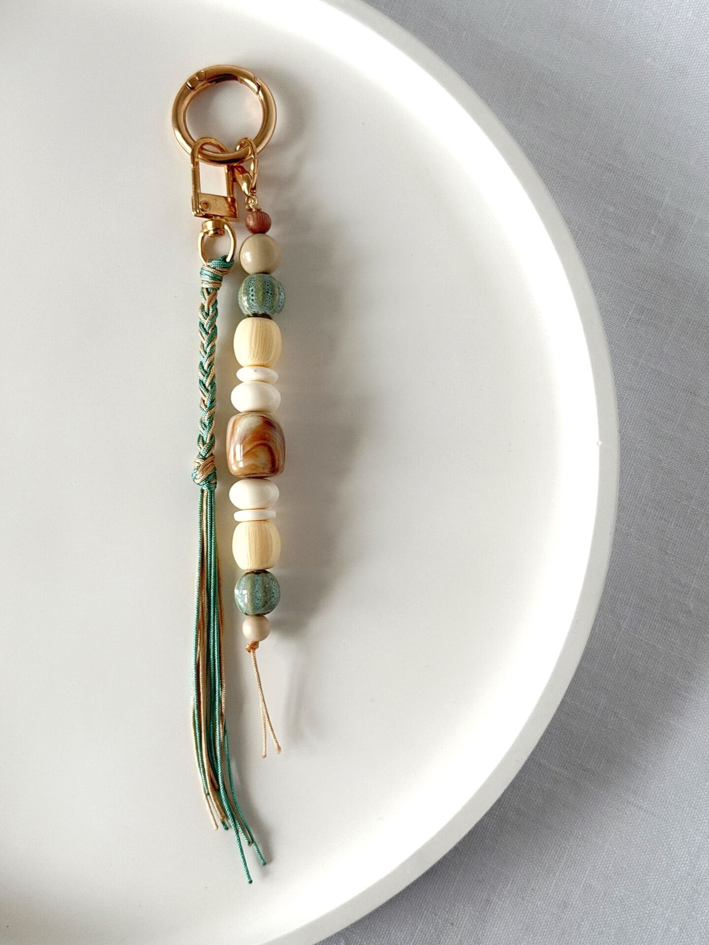 golden carabiner bag charm with blue and beige chunky beads with a braided nylon cord tassel in aqua and beige displayed on a white platter