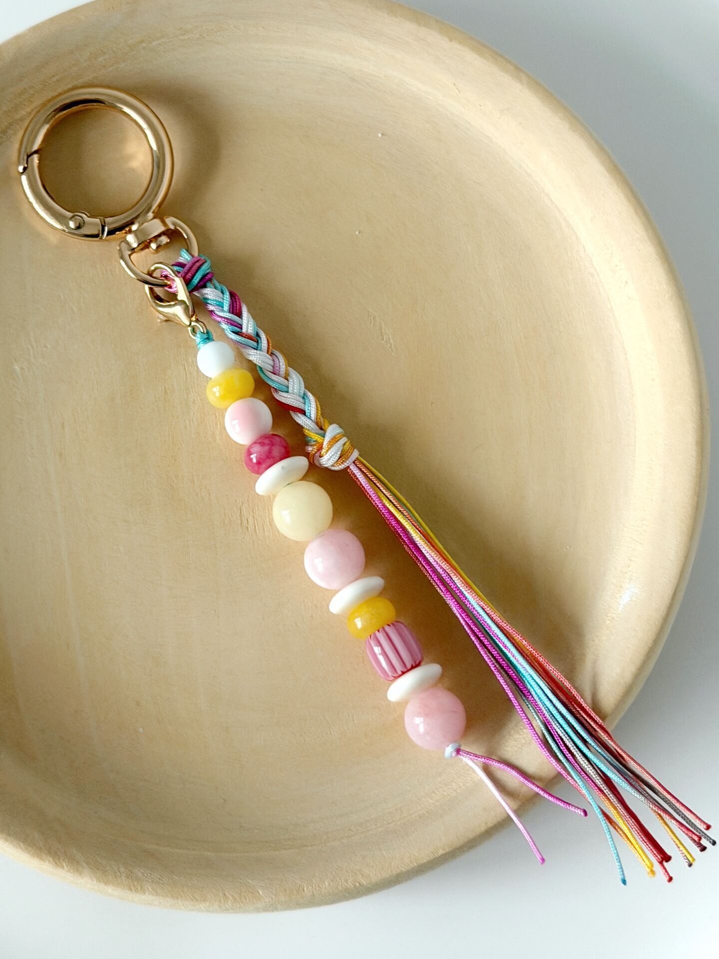 beaded bag charm with red, yellow, white and pink beads with a rainbow coloured nylon braided tassel displayed on a wooden plate