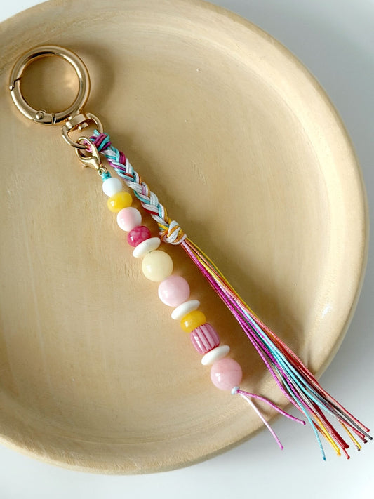 beaded bag charm with red, yellow, white and pink beads with a rainbow coloured nylon braided tassel displayed on a wooden plate