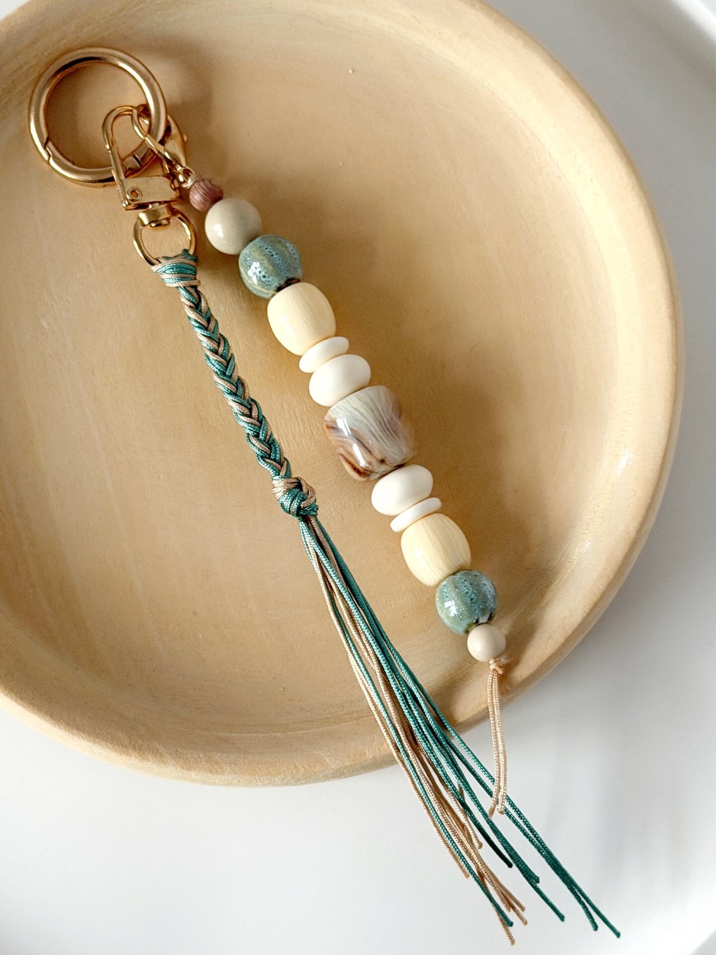 golden carabiner bag charm with blue and beige chunky beads with a braided nylon cord tassel in aqua and beige displayed on a wooden plate