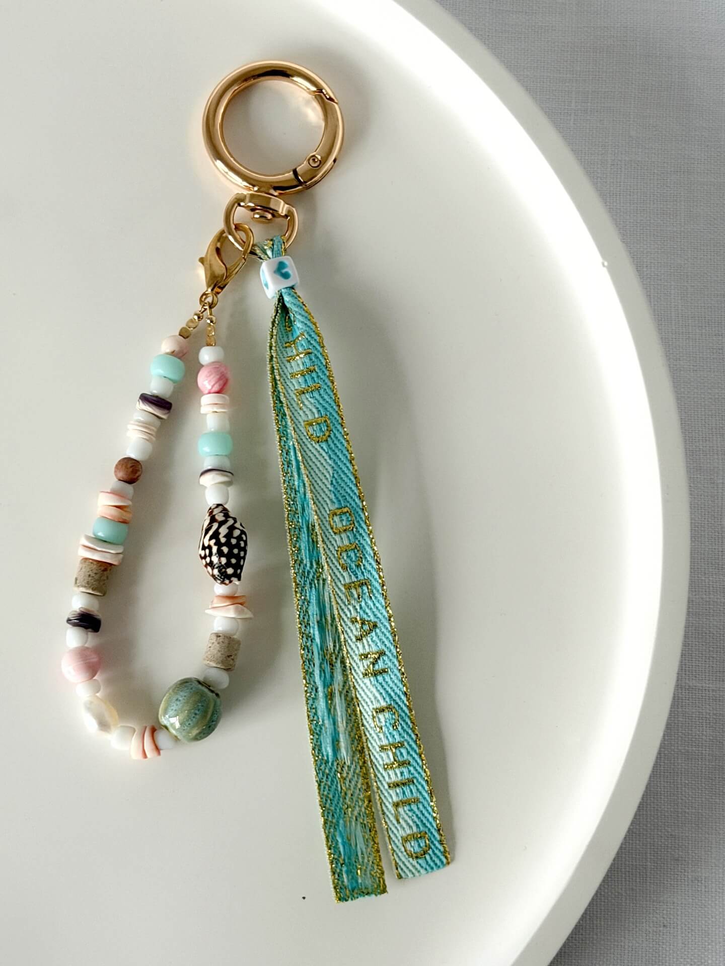 golden carabiner clasp bag charm with shell, glass, and wood beads with a blue and gold embroidered ocean child ribbon tassel