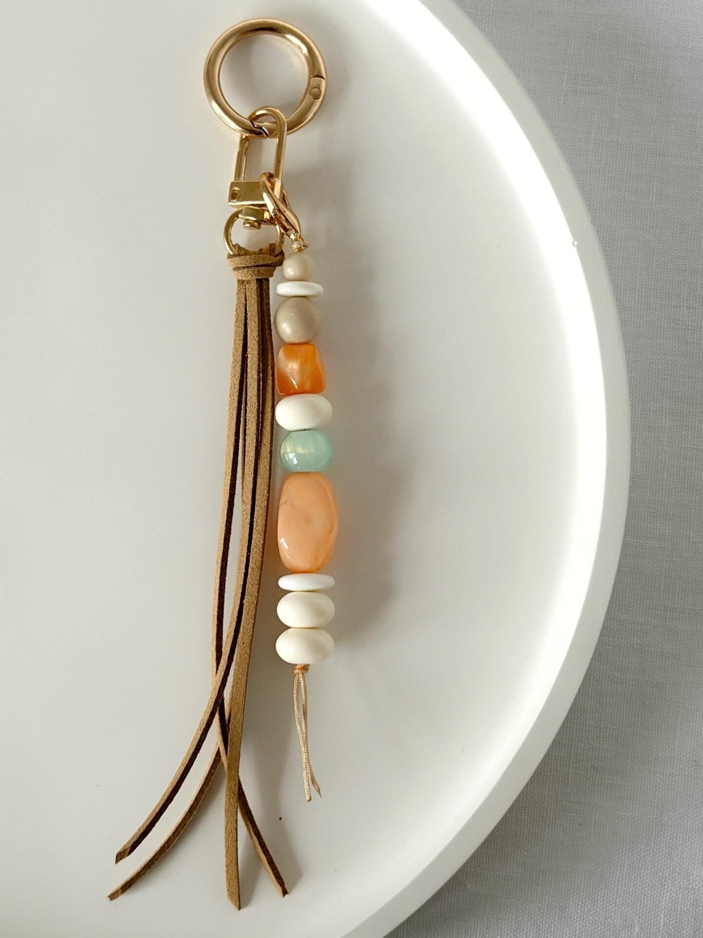 gold bag charm or key ring with long beige faux suede tassel and resin and stone beads in beige, orange, cream and aqua displayed on a white background