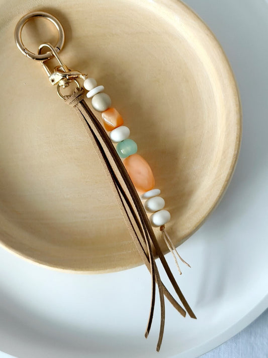 gold bag charm or key ring with long beige faux suede tassel and resin and stone beads in beige, orange, cream and aqua displayed against a wooden plate