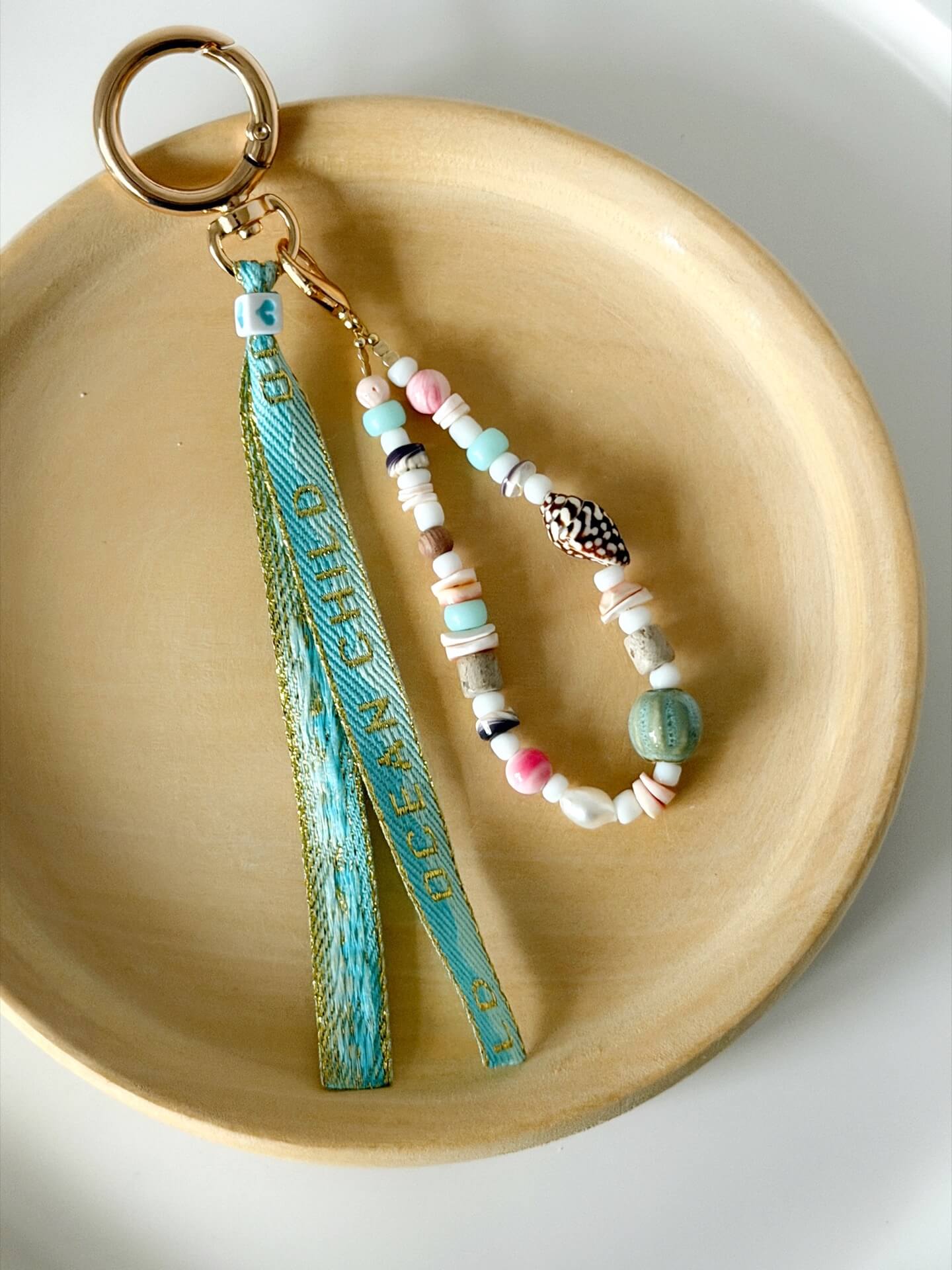 golden carabiner clasp bag charm with shell, glass, and wood beads with a blue and gold embroidered ocean child ribbon tassel displayed on a wooden plate