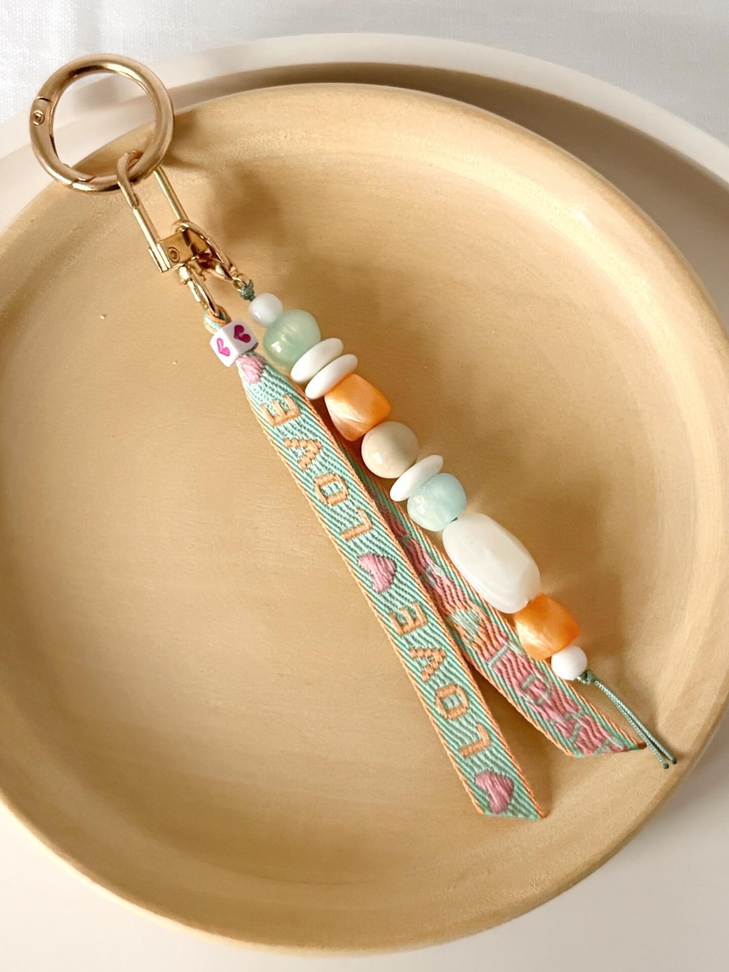 beaded bag charm with a love embroidered ribbon and aqua, white and orange chunky resin beaded on a gold clasp displayed against a wooden plate