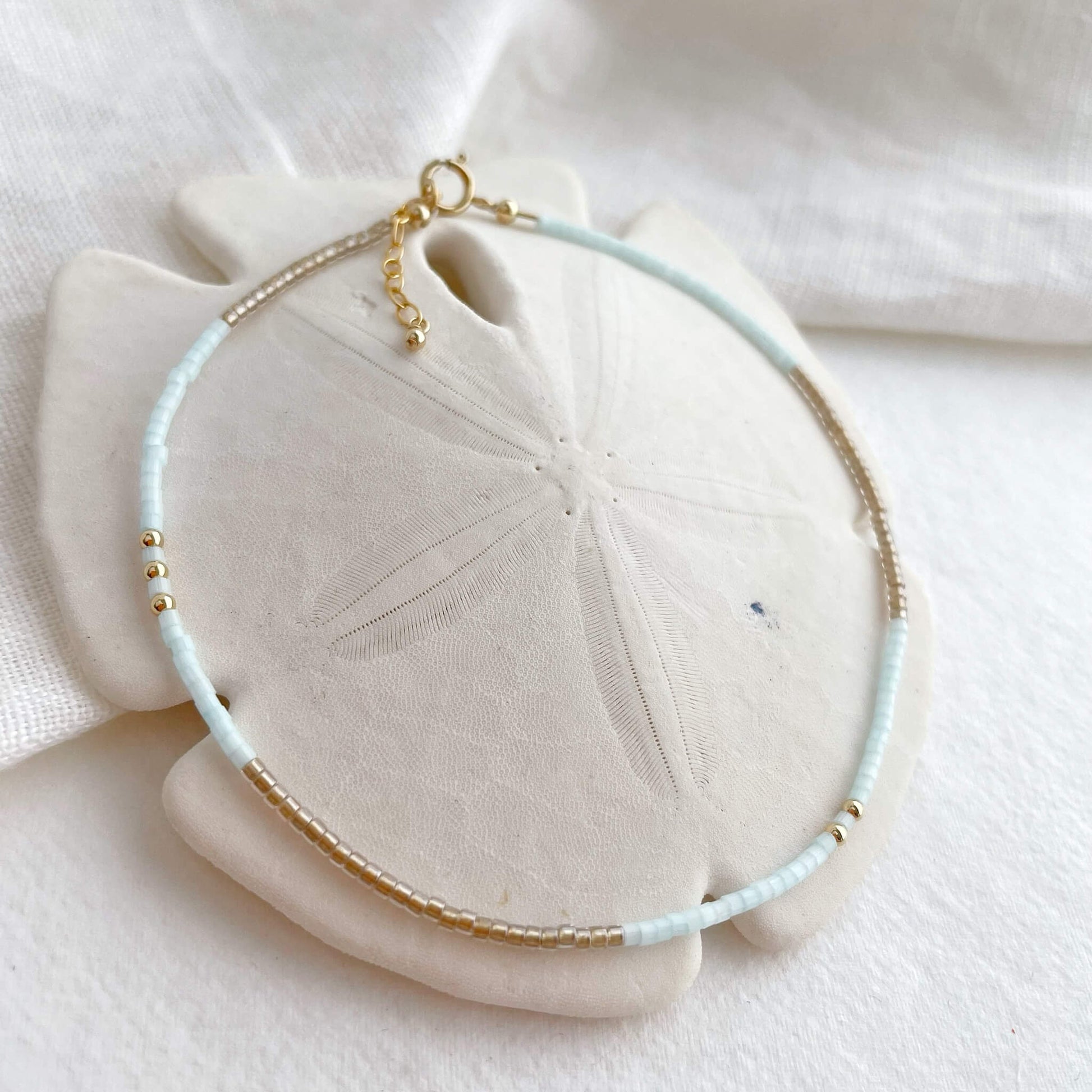 Ice Gold anklet with ice blue and golden Japanese glass beads and gold fill accents shown displayed draped over a white sand dollar