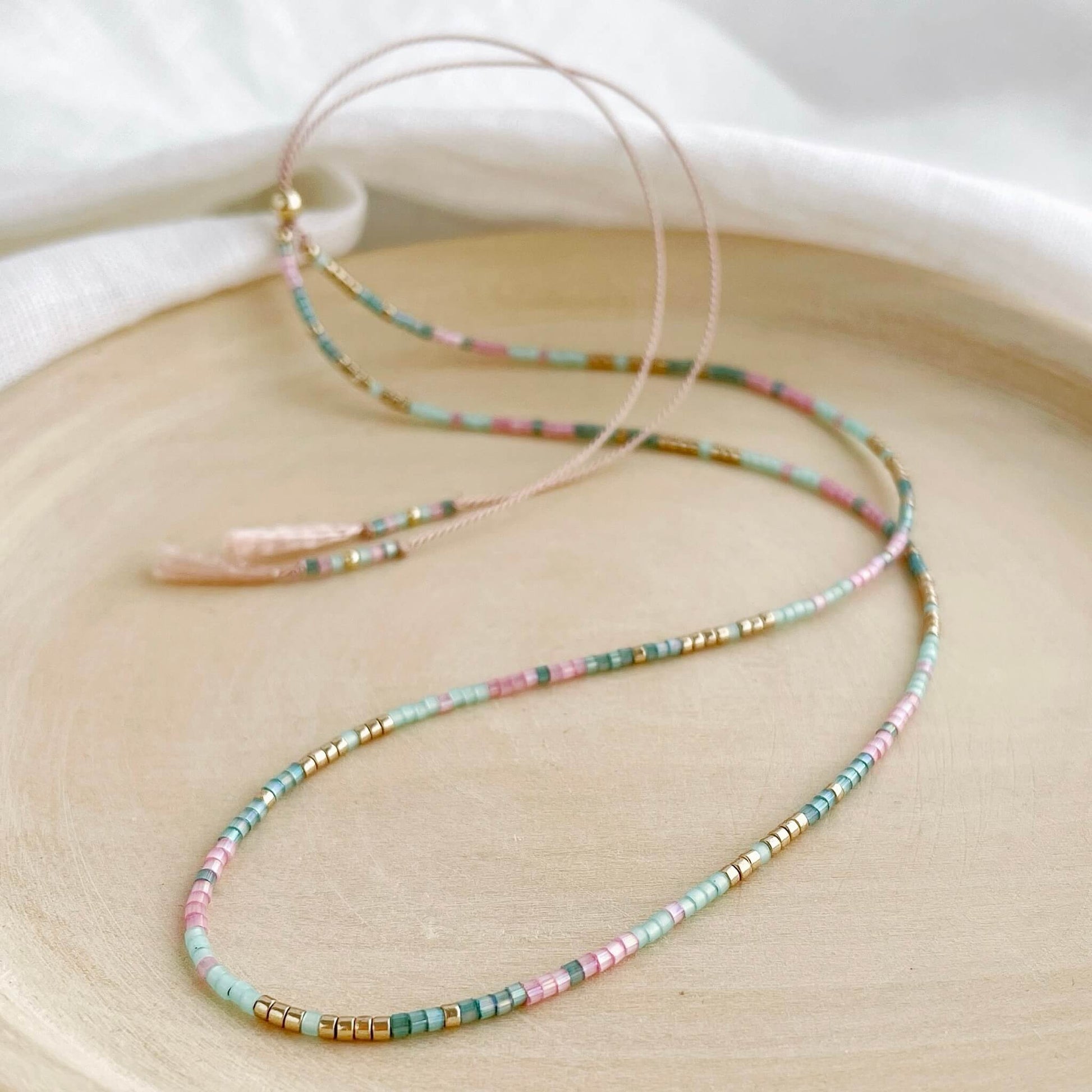 Pink and green silk beaded necklace with gold accents on a pale pink silk cord displayed on a wooden plate