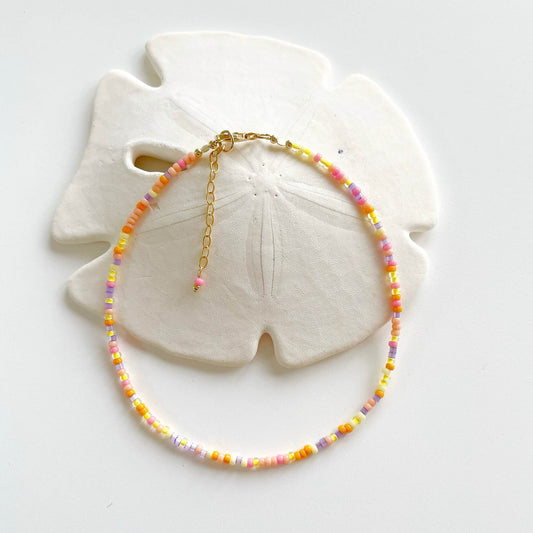 Sunset inspired anklet with pink, orange, yellow and lilac beads and gold fill accents displayed draped over a sand dollar
