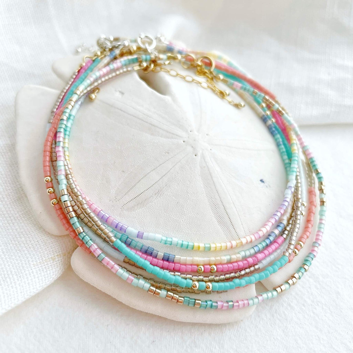 a mixture of 7 dainty multicoloured beaded anklets wrapped together over a white sand dollar