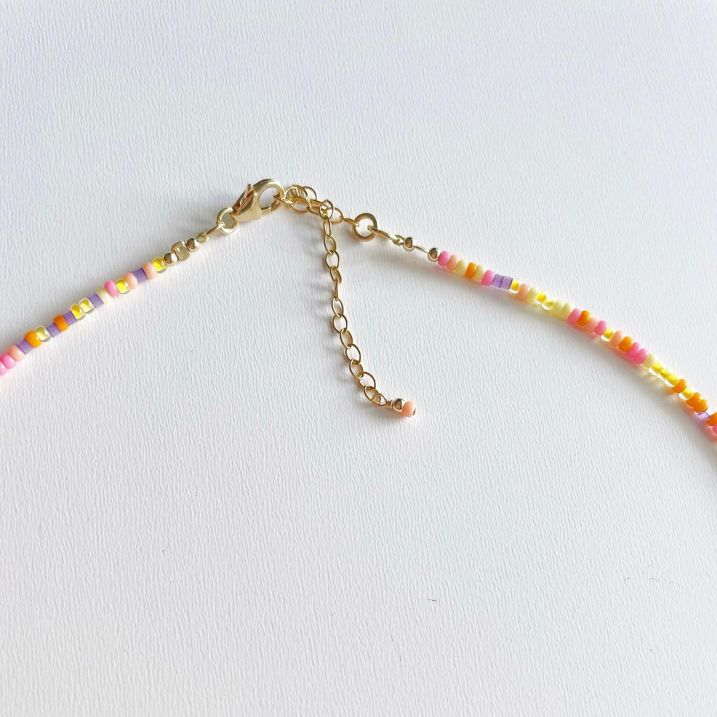 section of a beaded anklet with tiny pink, orange, peach, yellow and lilac beads showing the gold lobster clasp and extension chain