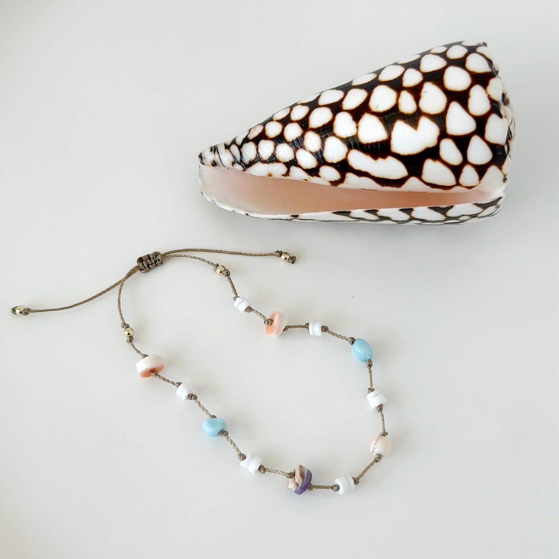 Beige knotted nylon cord bracelet with Larimar gemstone, pink conch shell, orange shell and purple wampum shell beads on a neutral surface with a shell