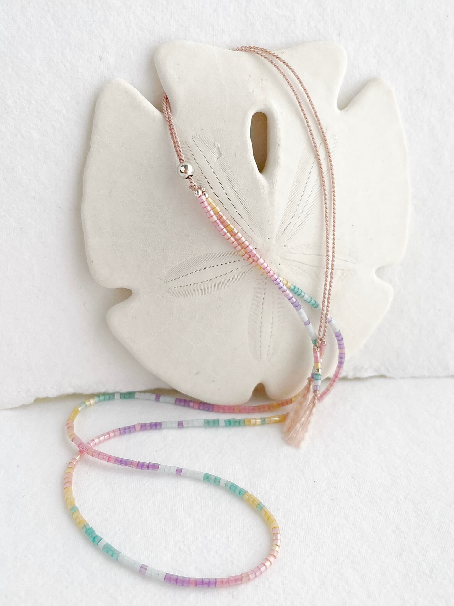 dainty Rainbow glass bead necklace on pale pink silk cord with silver clasp displayed draped over a sand dollar