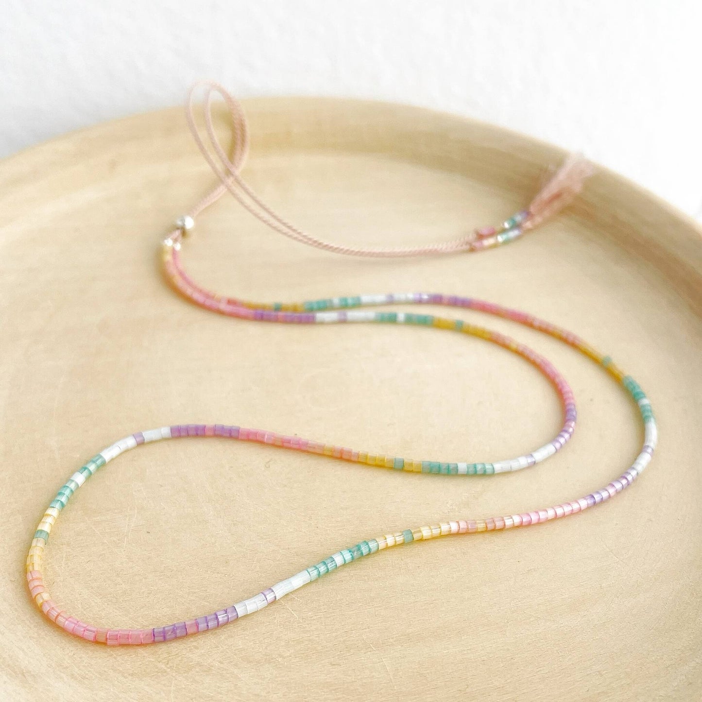 Rainbow glass bead necklace on pale pink silk cord with silver clasp displayed on a wooden plate