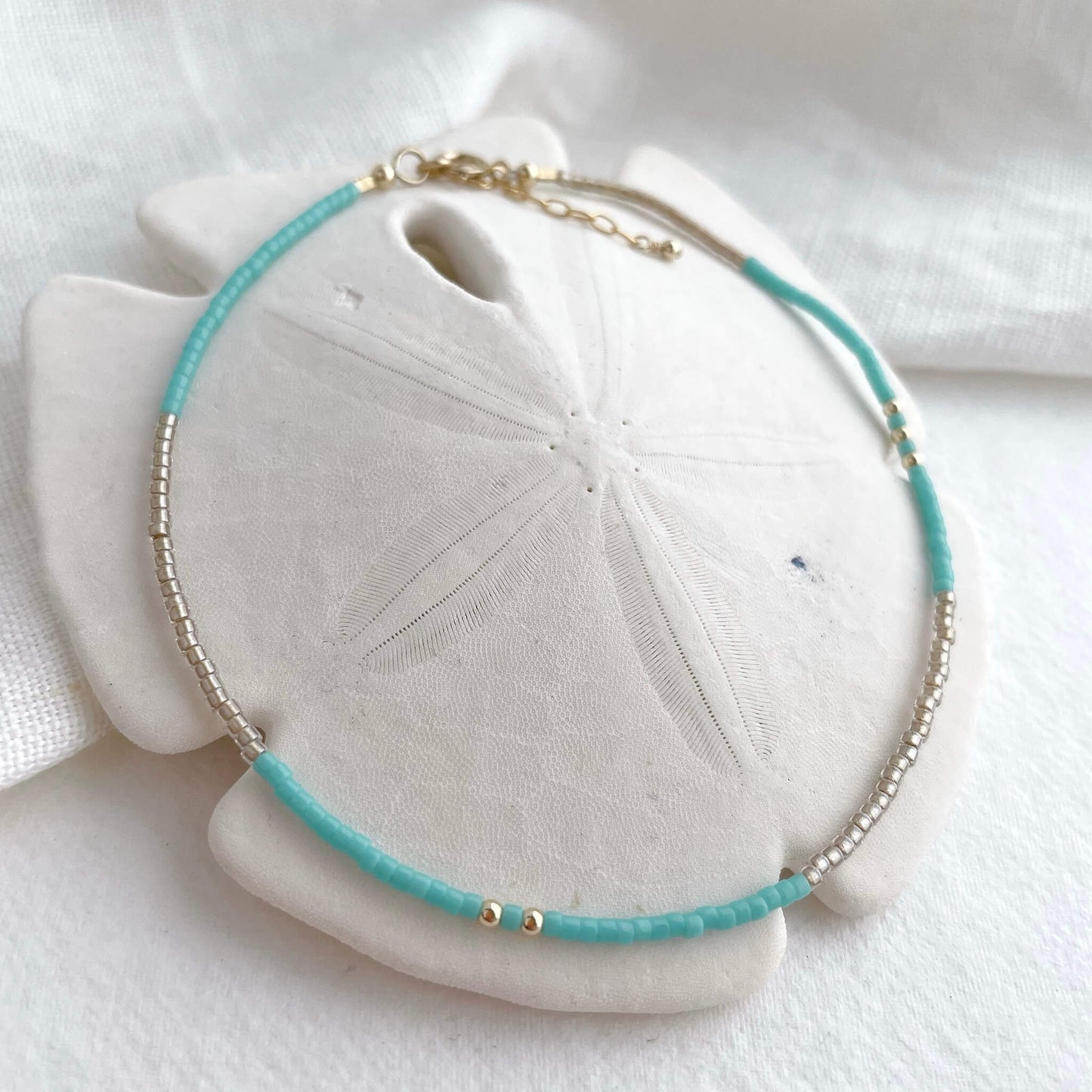 fistral anklet with turquoise and golden Japanese glass beads and gold fill accents displayed draped over a white sand dollar