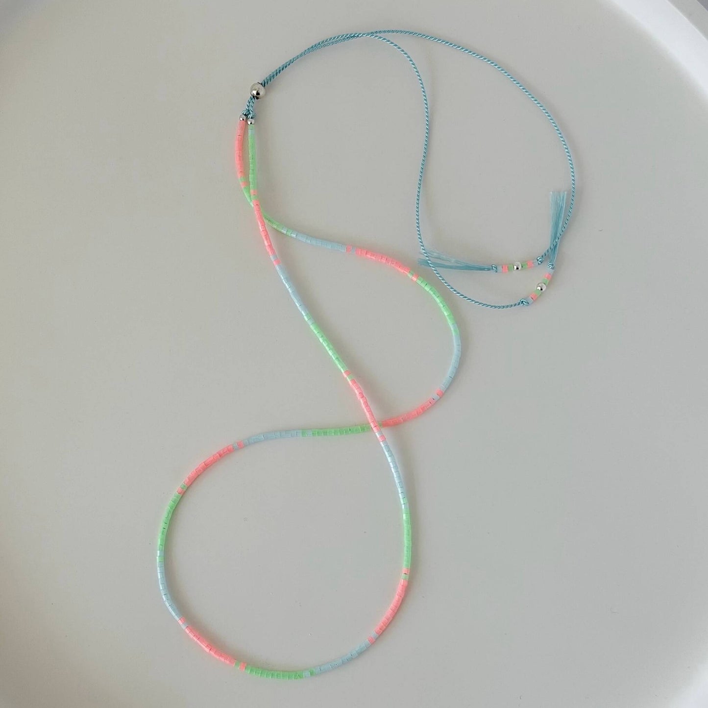 Dainty mint green, pink and pale blue glass bead necklace with sterling silver sliding clasp on a pale blue silk cord on a  white flat lay surface