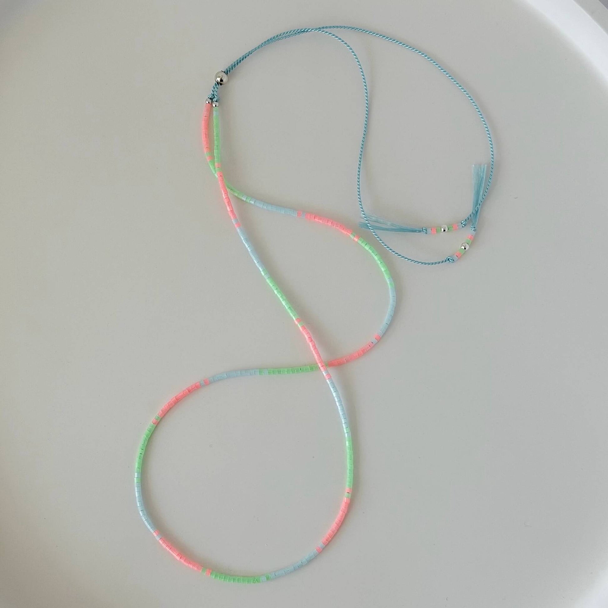 Dainty mint green, pink and pale blue glass bead necklace with sterling silver sliding clasp on a pale blue silk cord on a  white flat lay surface