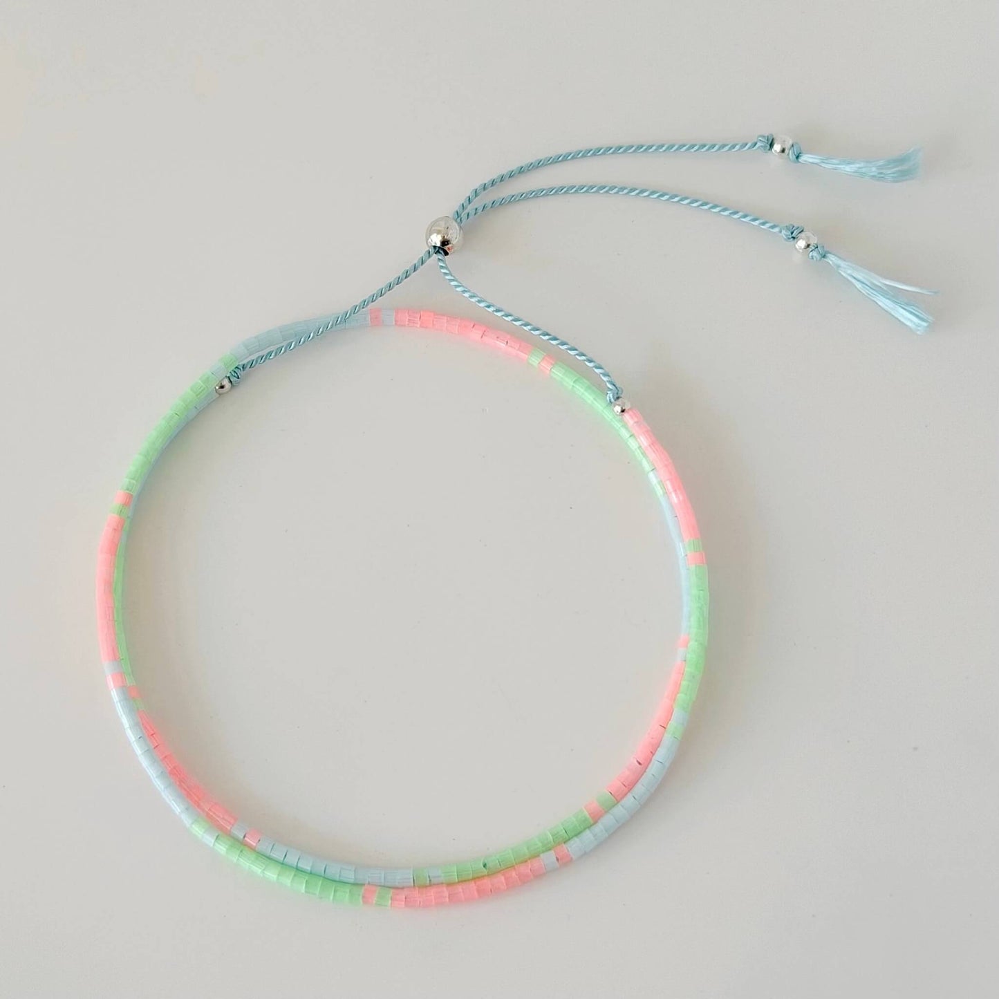 Dainty double wrap green, pink and pale blue glass bead bracelet with sterling silver sliding clasp on a pale blue silk cord against a  neutral surface